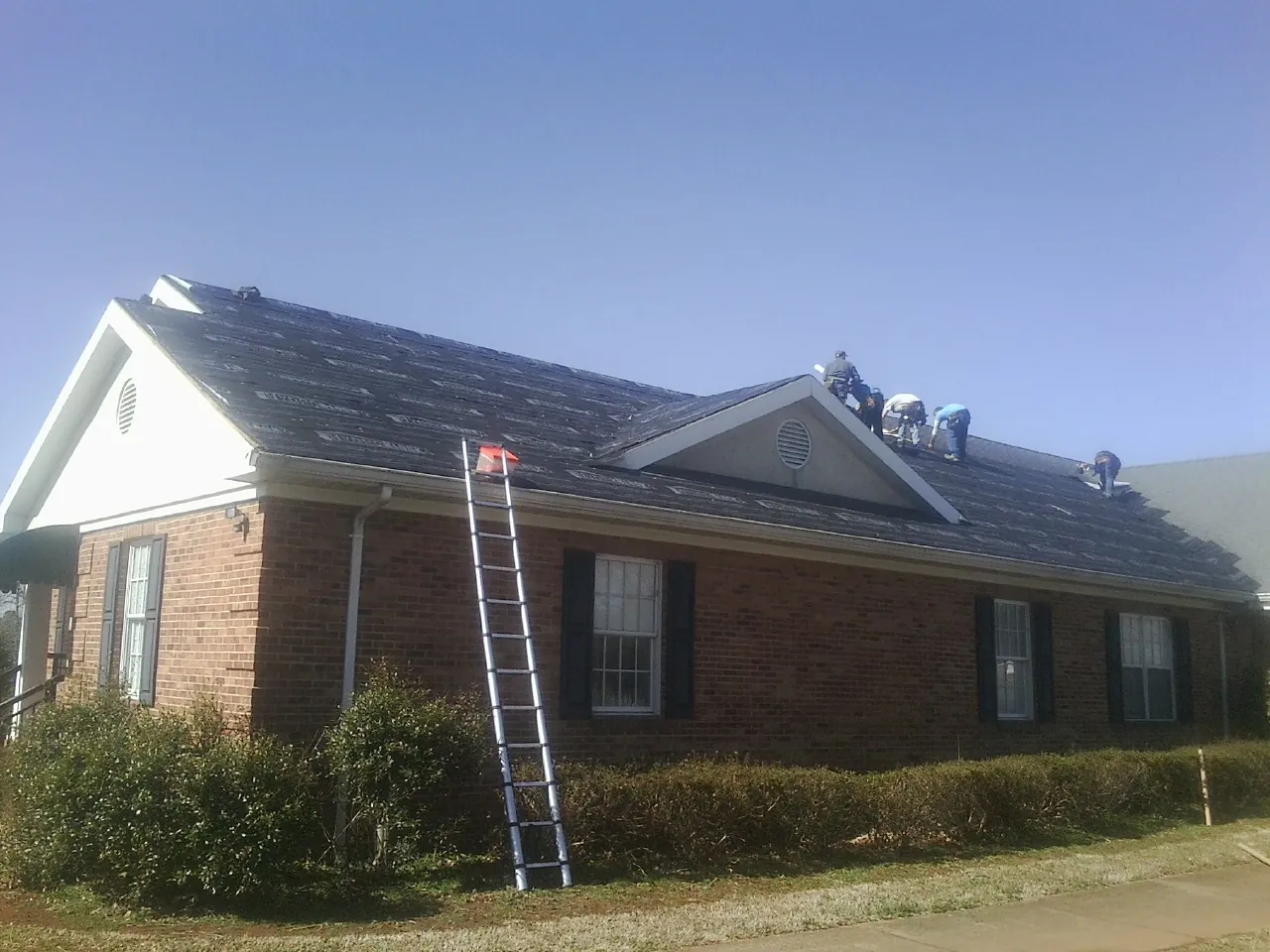 Crew tearing shingles off a brick ranch roof with a ladder set — Metal Roof Repair work by our crew in Nisswa