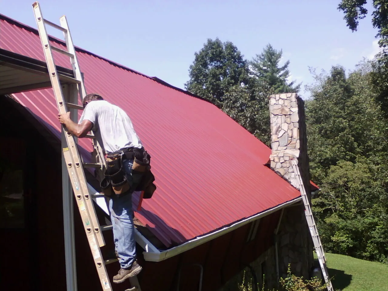 Roofer on a ladder beside a red metal roof and stone chimney — Soffit & Fascia Repair by Roofing Brothers in Nisswa